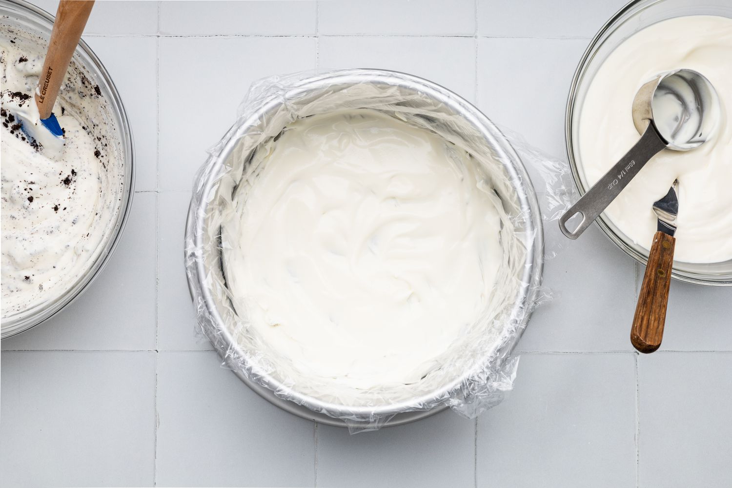 Assembling First Layer of Oreo Ice Cream Cake (L to R): Bowl of Cookies and Cream Whipped Layer, Plastic Wrapped Circular Cake Pan With No Churn Ice Cream, and a Bowl With More No Churn Ice Cream With a Measuring Cup and an Offset Spatula