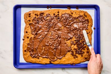 baked flattened cookie on making tray, chocolate chips scattered on top, hand spreading melting chocolate with a small spatula