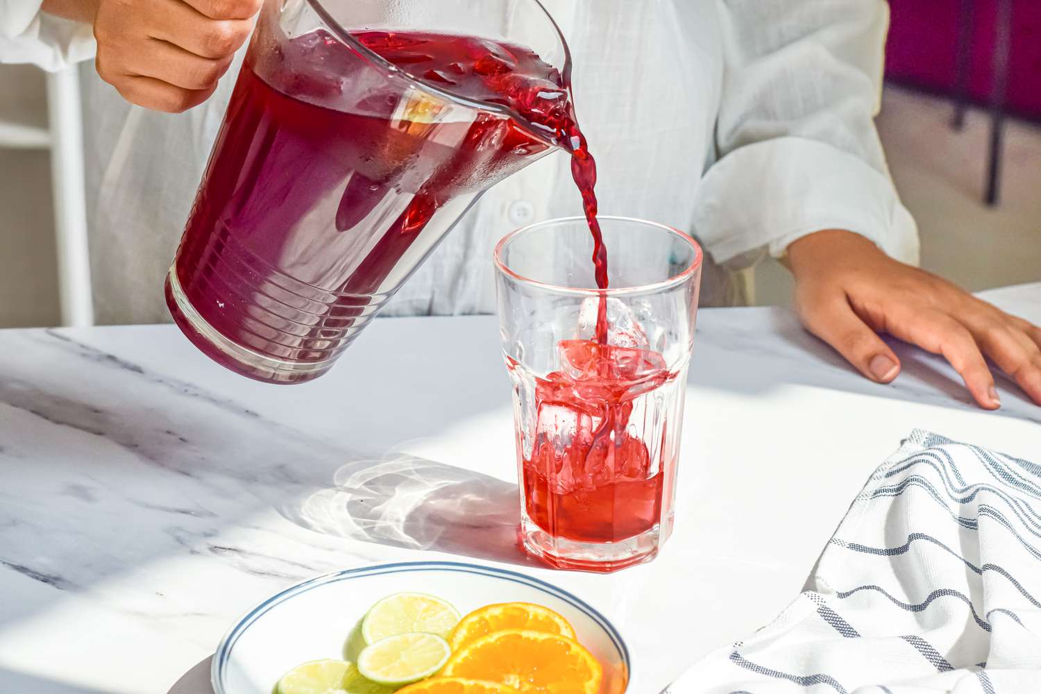 Agua de Jamaica (Hibiscus Tea) Poured into a Glass of Ice From a Pitcher. On the Counter with the Glass is a Plate with Lime and Orange Slices and a Kitchen Towel.