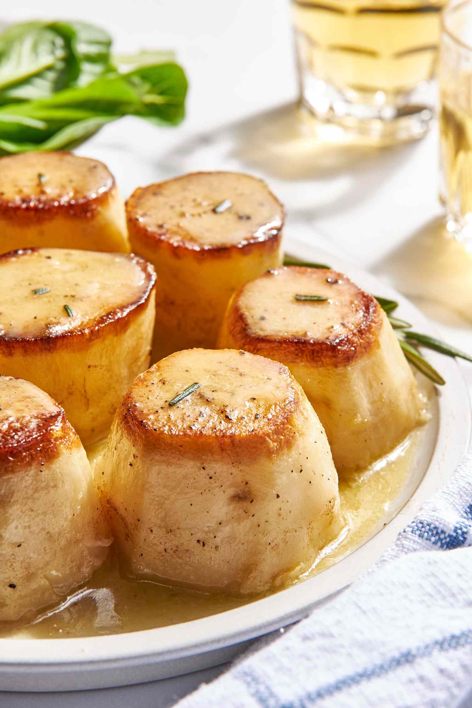 Fondant potatoes on a plate, and in the surroundings, a kitchen towel, two glasses, and a bowl of salad greens