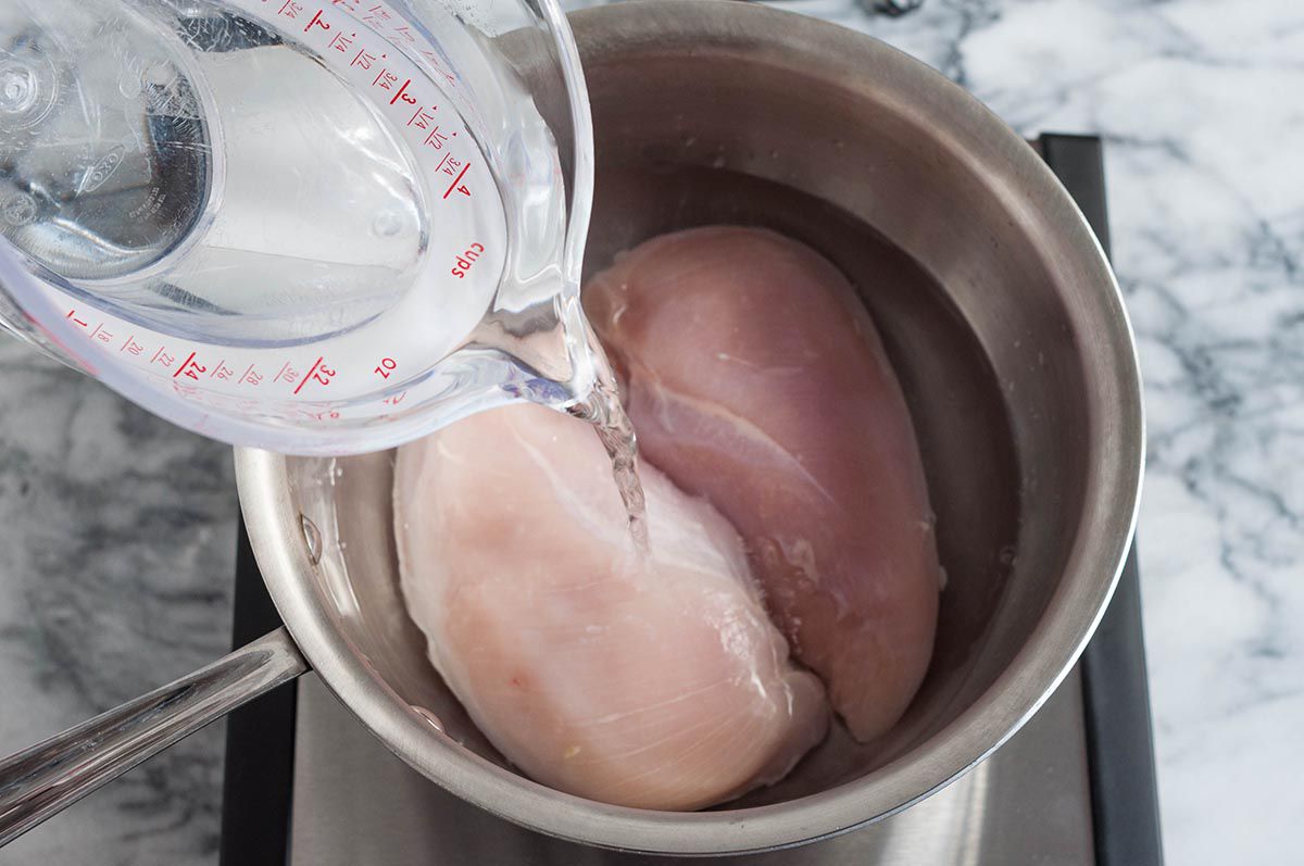 A hand pouring water into a pot with two chicken breasts inside