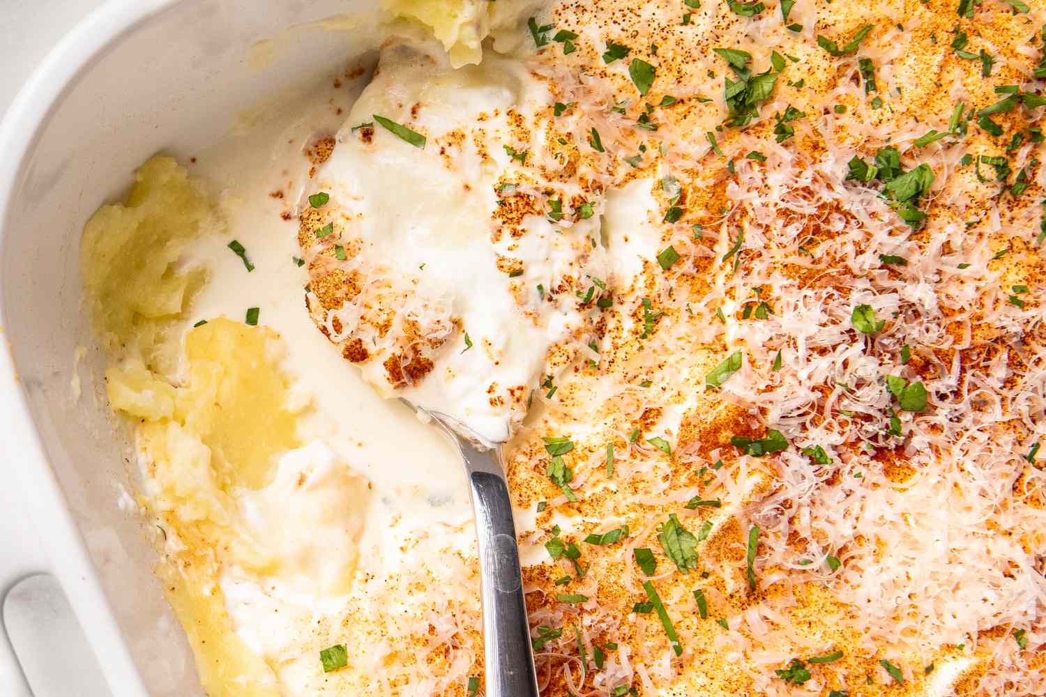 Casserole dish with a creamy layered casserole topped with grated cheese and parsley part of the dish served with a spoon
