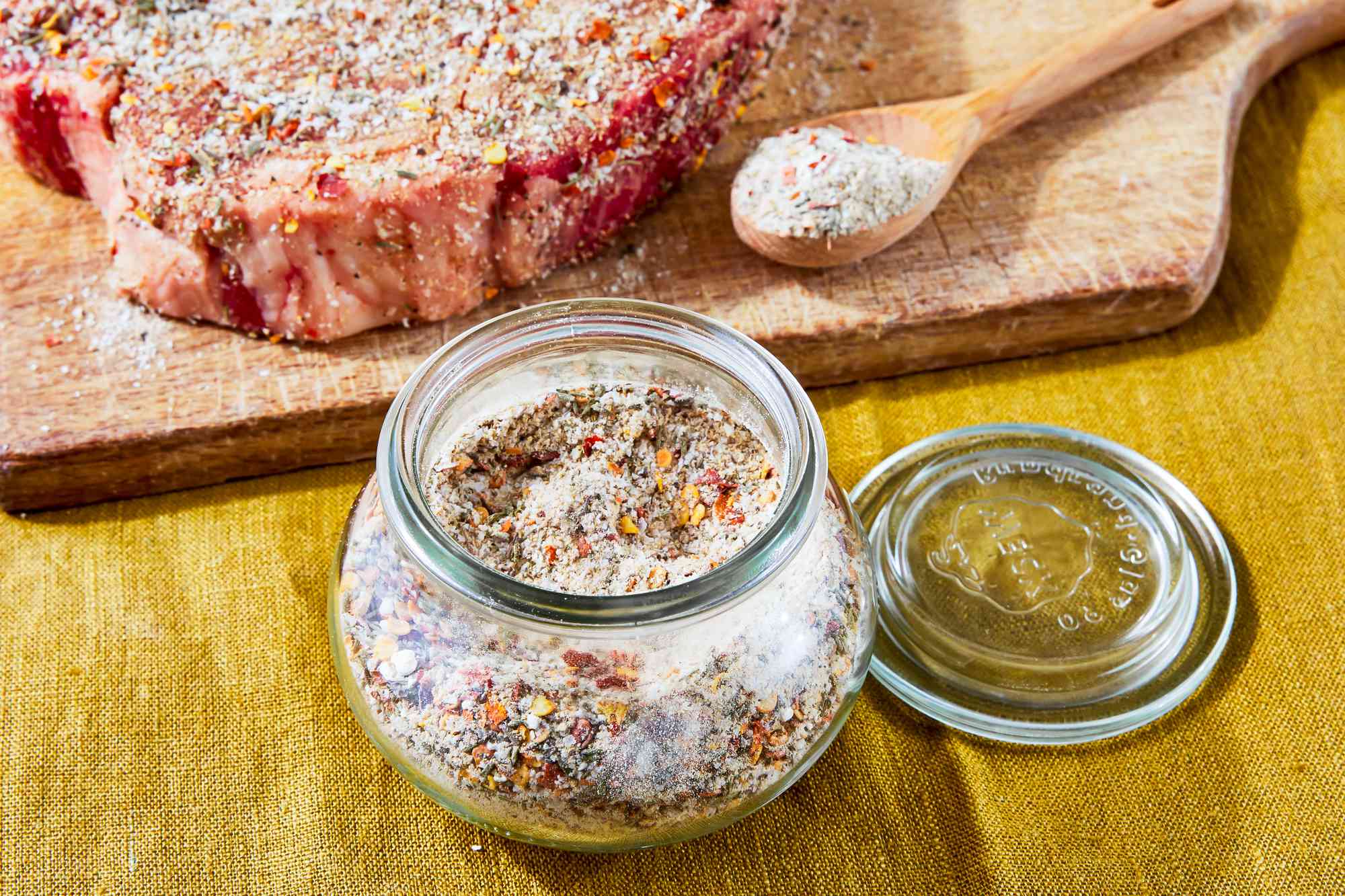 Jar of Steak Seasoning on the Counter, and in the Background, a Raw Seasoned Steak and a Spoon With More Seasoning on a Cutting Board