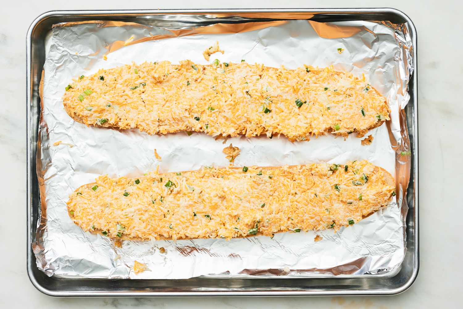 Garlic Butter and Cheese Mixture Spread on Two Halves of a French Loaf on an Aluminum Lined Baking Sheet 