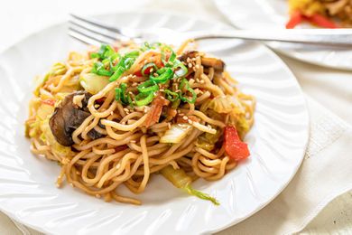 Side view of a white plate and fork with homemade vegetable lo mein. A fork is on the plate and a second plate is partially visible in the upper right hand corner.