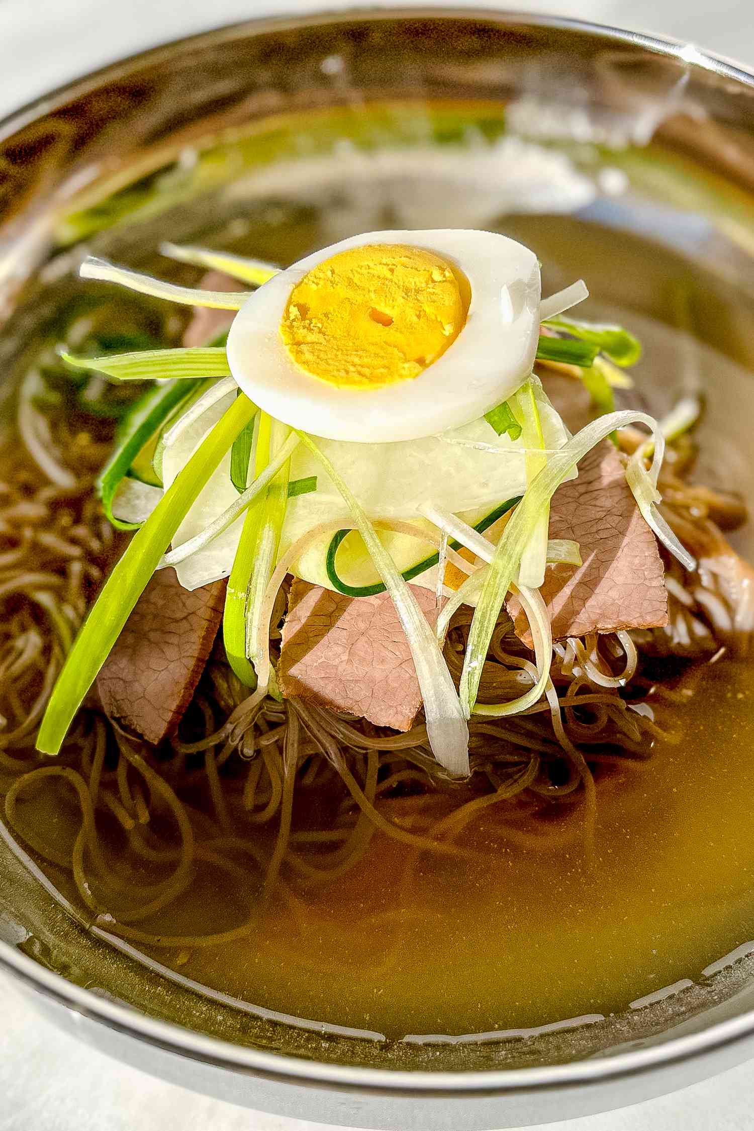 Bowl of Naengmyeon Topped With a Halved Hard Boiled Egg, Cucumber Ribbons, and Julienned Green Onions