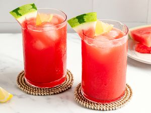 Two glasses of watermelemonade with ice and fruit garnishes placed on coasters on a surface