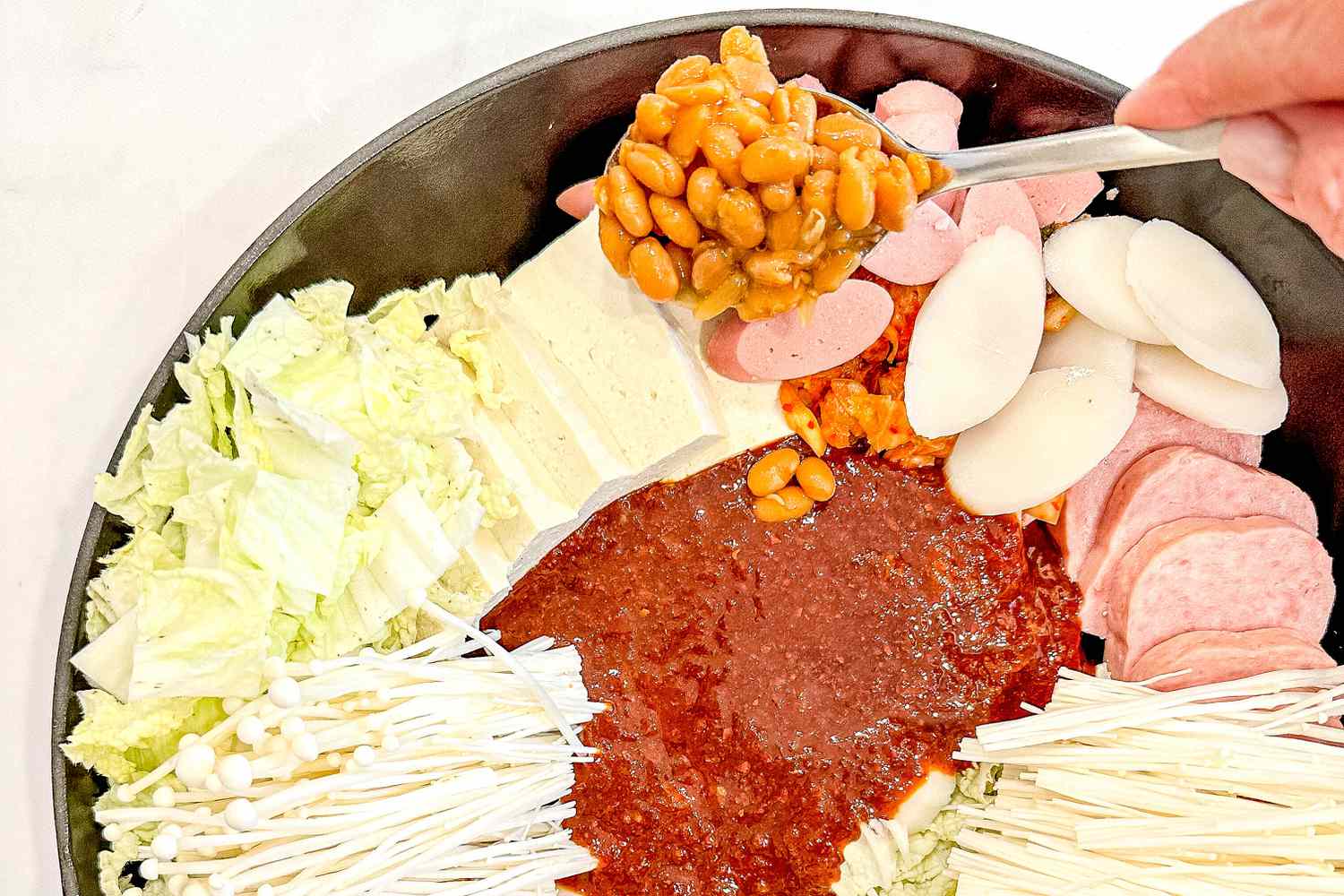 Canned baked beans added to pot of nicely organized budae jjigae ingredients 