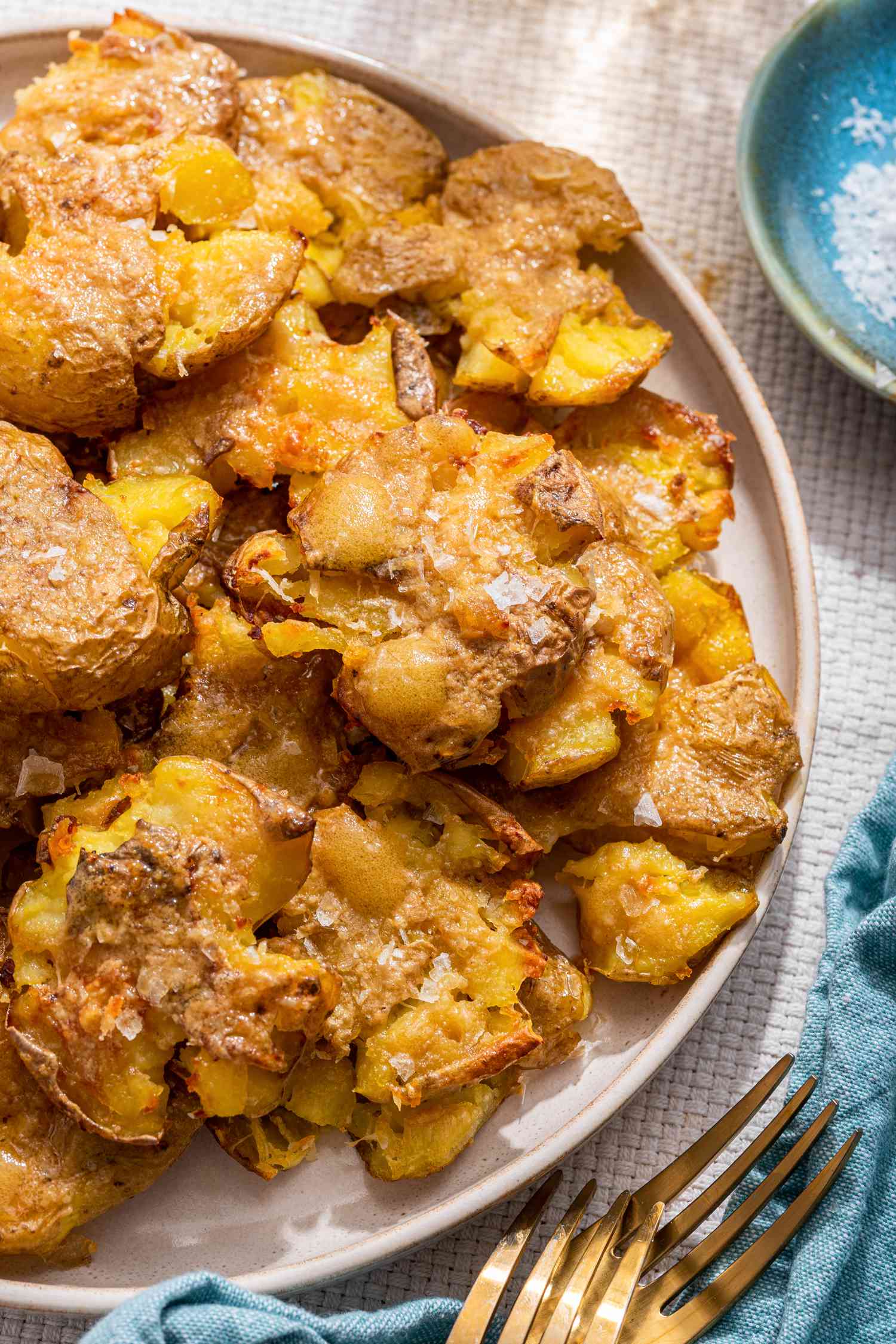 Crash hot potatoes on a platter next to a small saucer with flaky salt, utensils, and a kitchen towel