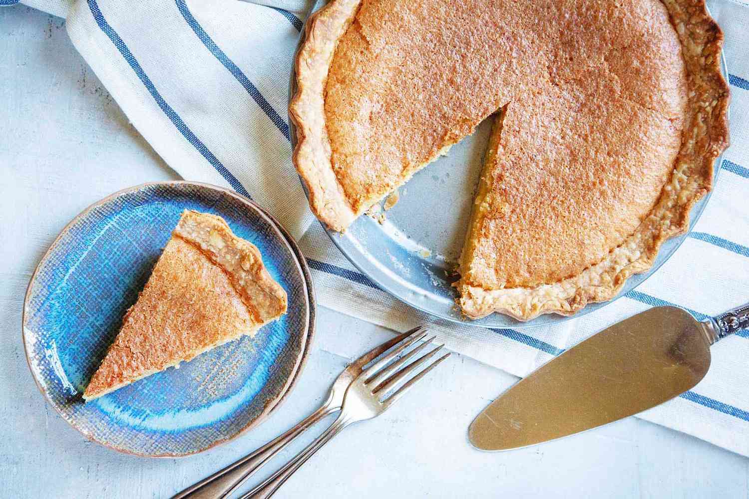 Overhead view of the best chess pie with a slice removed. The slice is on a blue plate with forks and a pie server to the right.