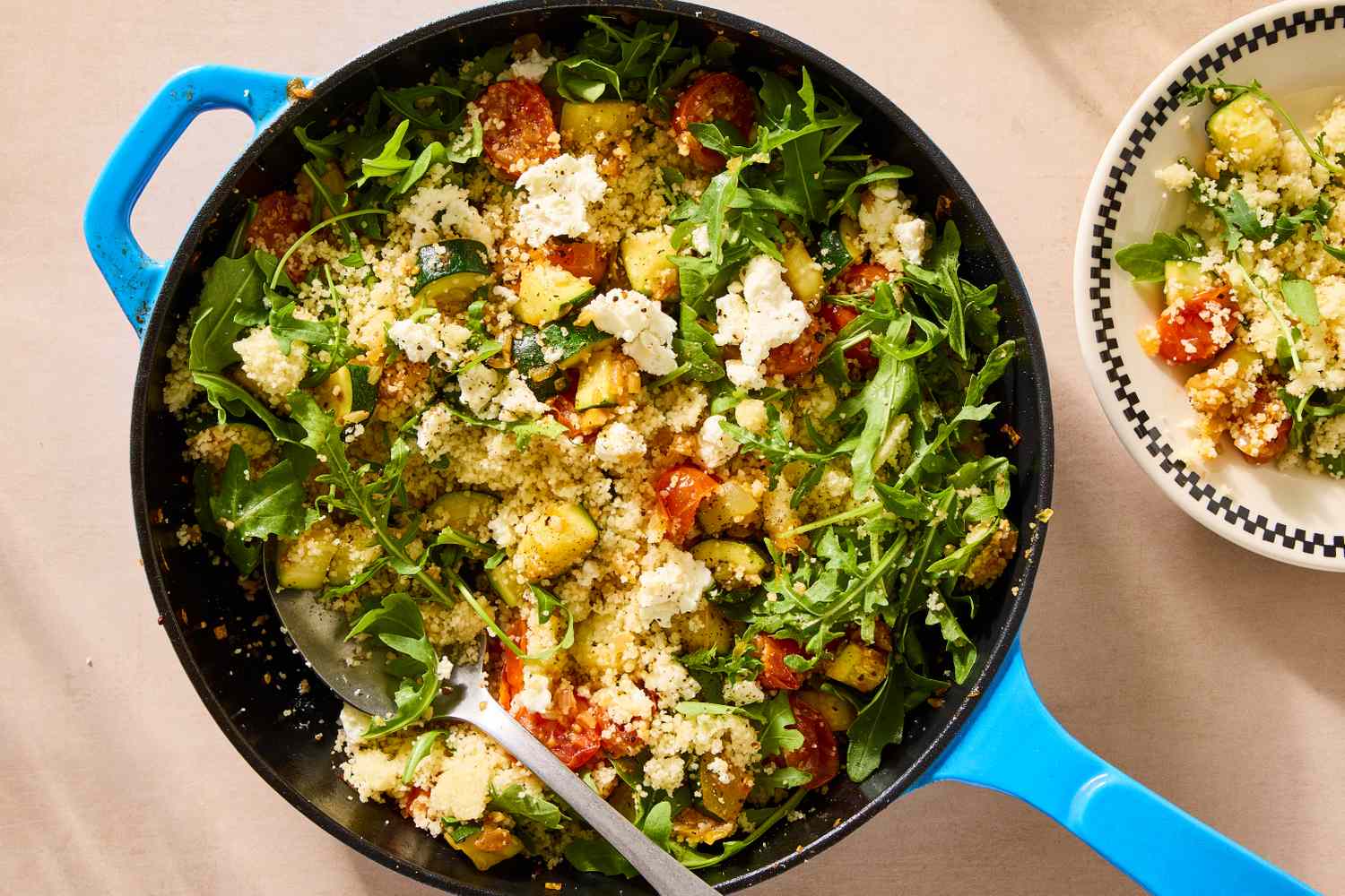 A skillet containing a dish of couscous mixed with vegetables and greens, topped with a visible spoon