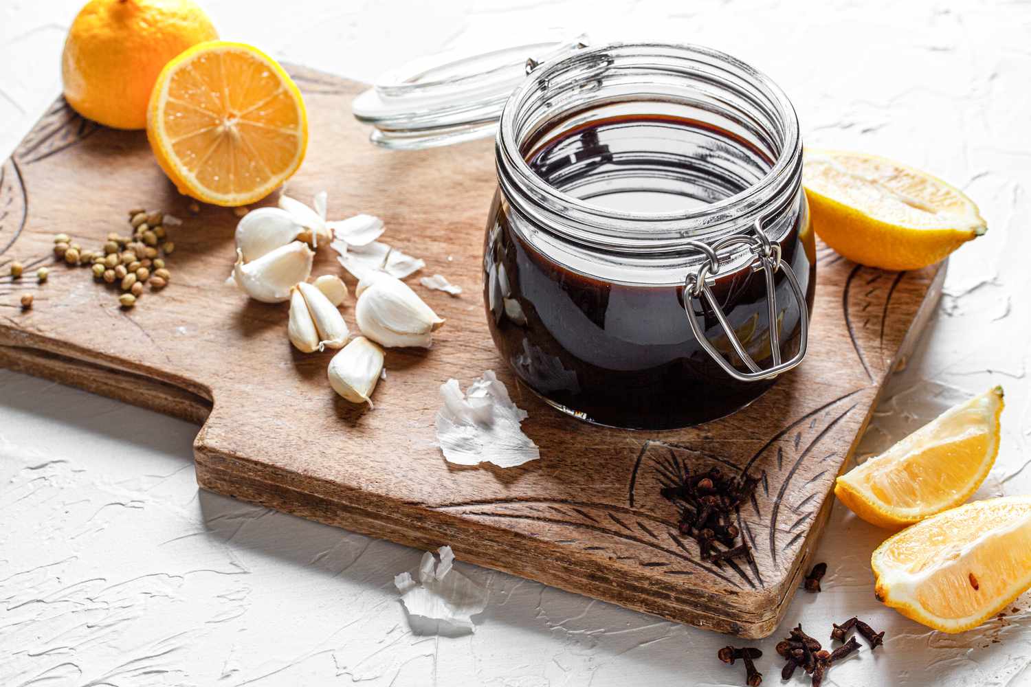 Jar of Homemade Worcestershire Sauce on a Decorative Wooden Board Covered in Ingredients (Halved Orange, Coriander Seeds, Garlic Cloves, Cloves, and Lemon Wedges)
