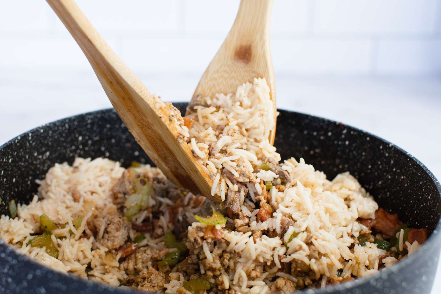 Two wooden utensils mix in rice to the meat and vegetable mixture to show how to make Cajun dirty rice.