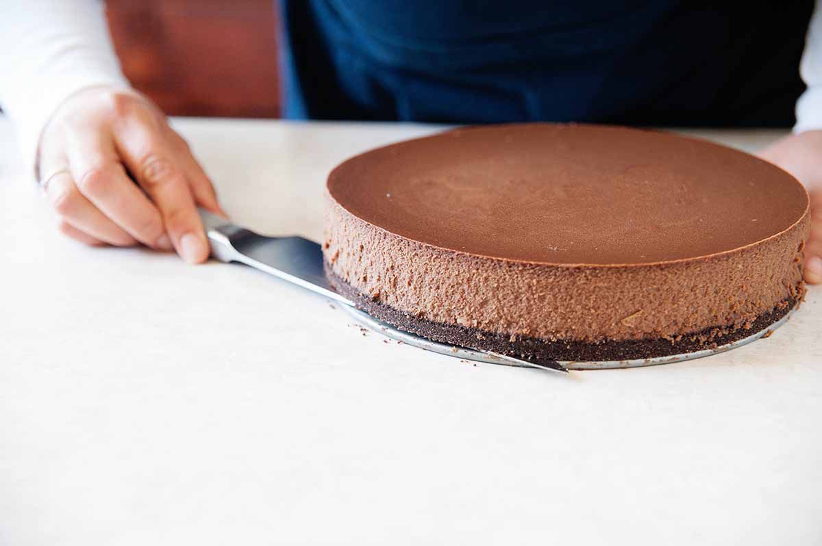 Sliding a knife between the bottom of a springform pan and the crust of a triple chocolate cheesecake.