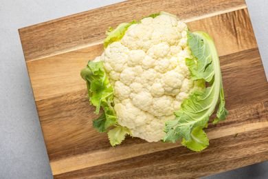 Whole cauliflower head on a wooden cutting board partially surrounded by green leaves