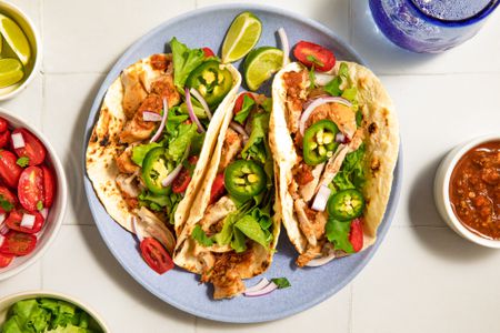 Plate of chicken tacos garnished with jalapeños, onions, and greens, served with lime wedges and salsa on the side