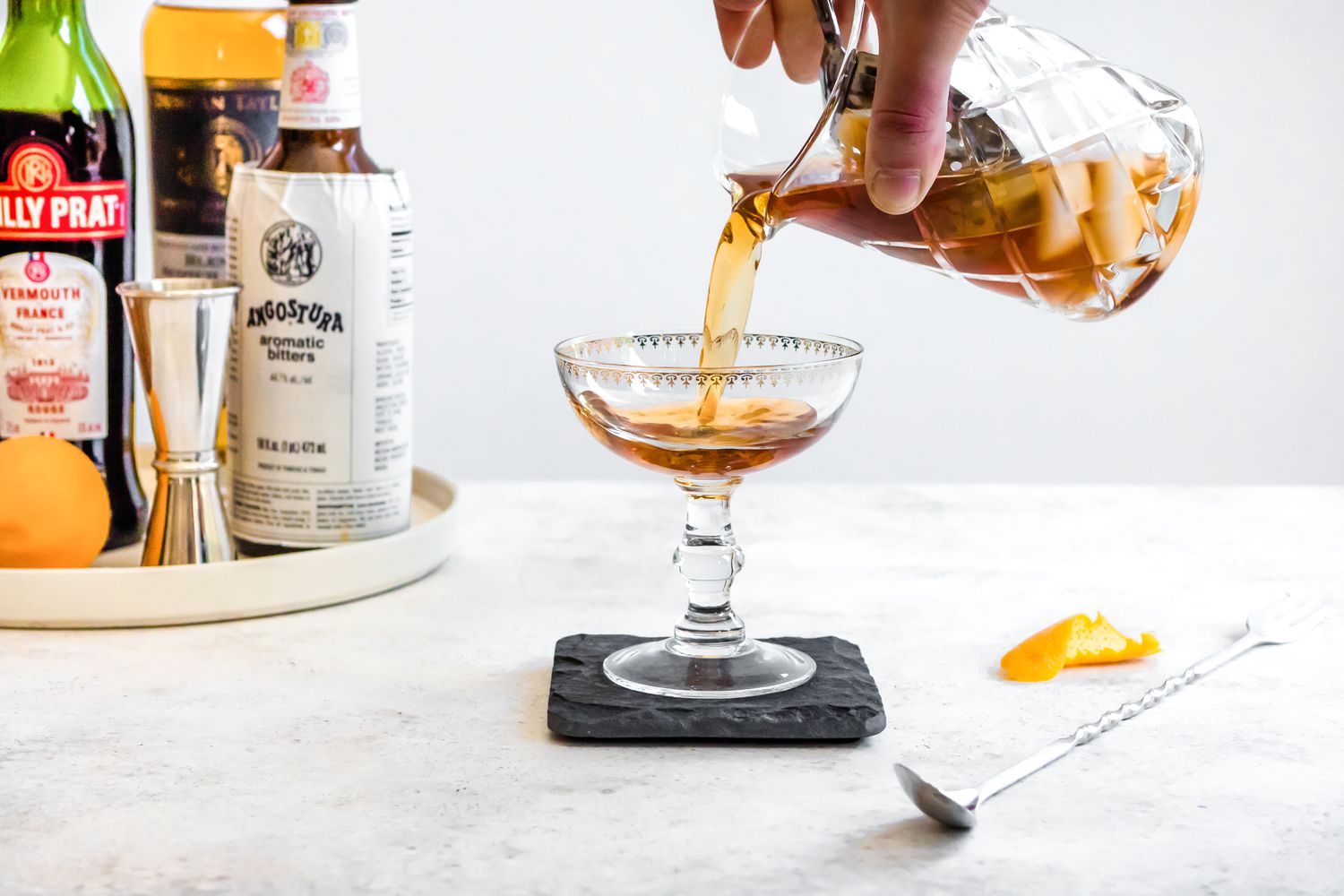 Pour Rob Roy into a Cocktail Glass from a Mixing Glass with a Tray of Recipe Ingredients (Vermouth, Whiskey, and Orange) in the Background