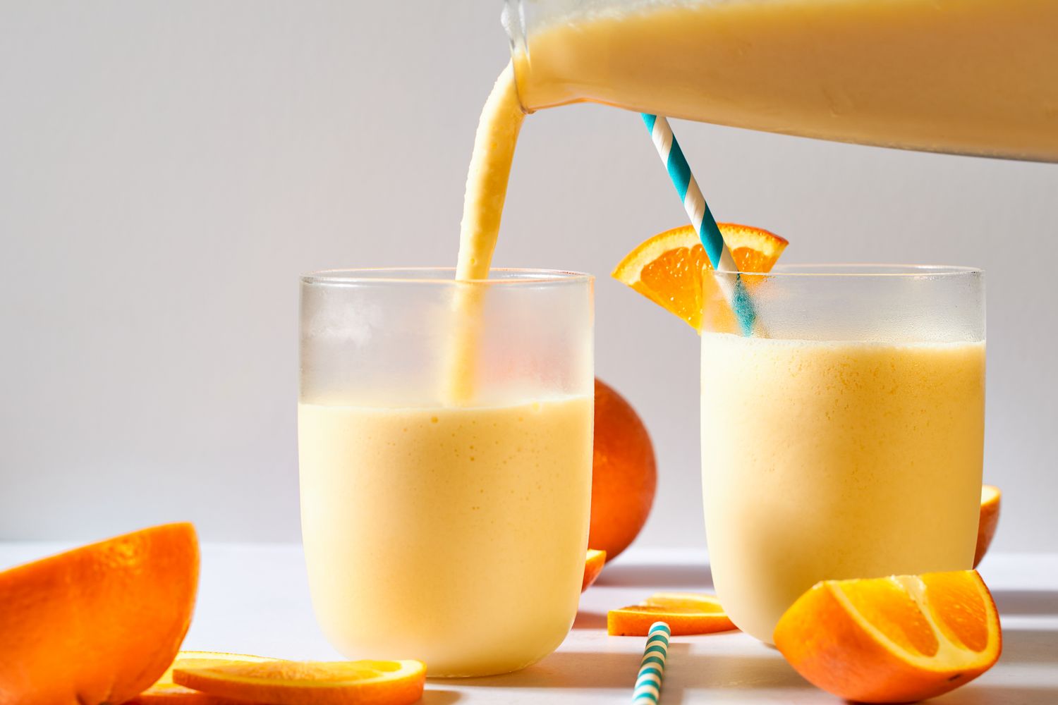 Two glasses of orange smoothie are served, with a blue-striped straw in one and orange slices on the rim and table