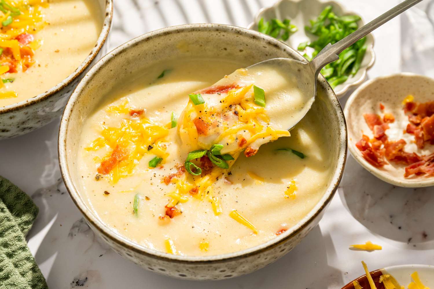 Closeup view of a bowl of potato soup topped with bacon, shredded cheese and green onions next to a bowls of bacon, green onions and shredded cheese