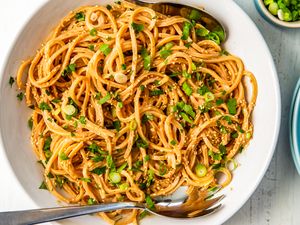 Peanut butter noodles garnished with sesame seeds and scallions in a bowl with serving utensils