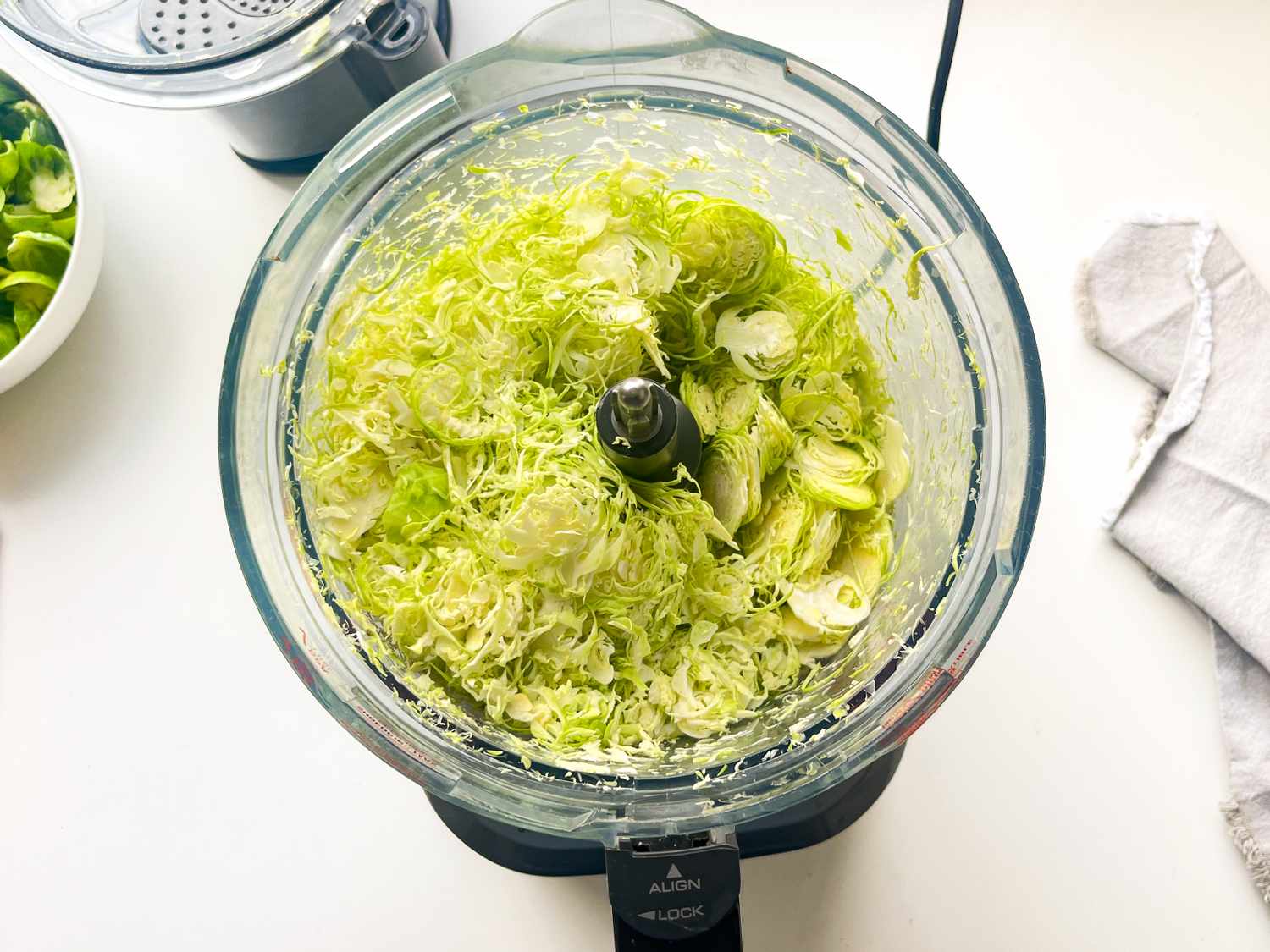 Shredded Brussels sprouts in a food processor bowl bowl of sprouts and kitchen towel nearby