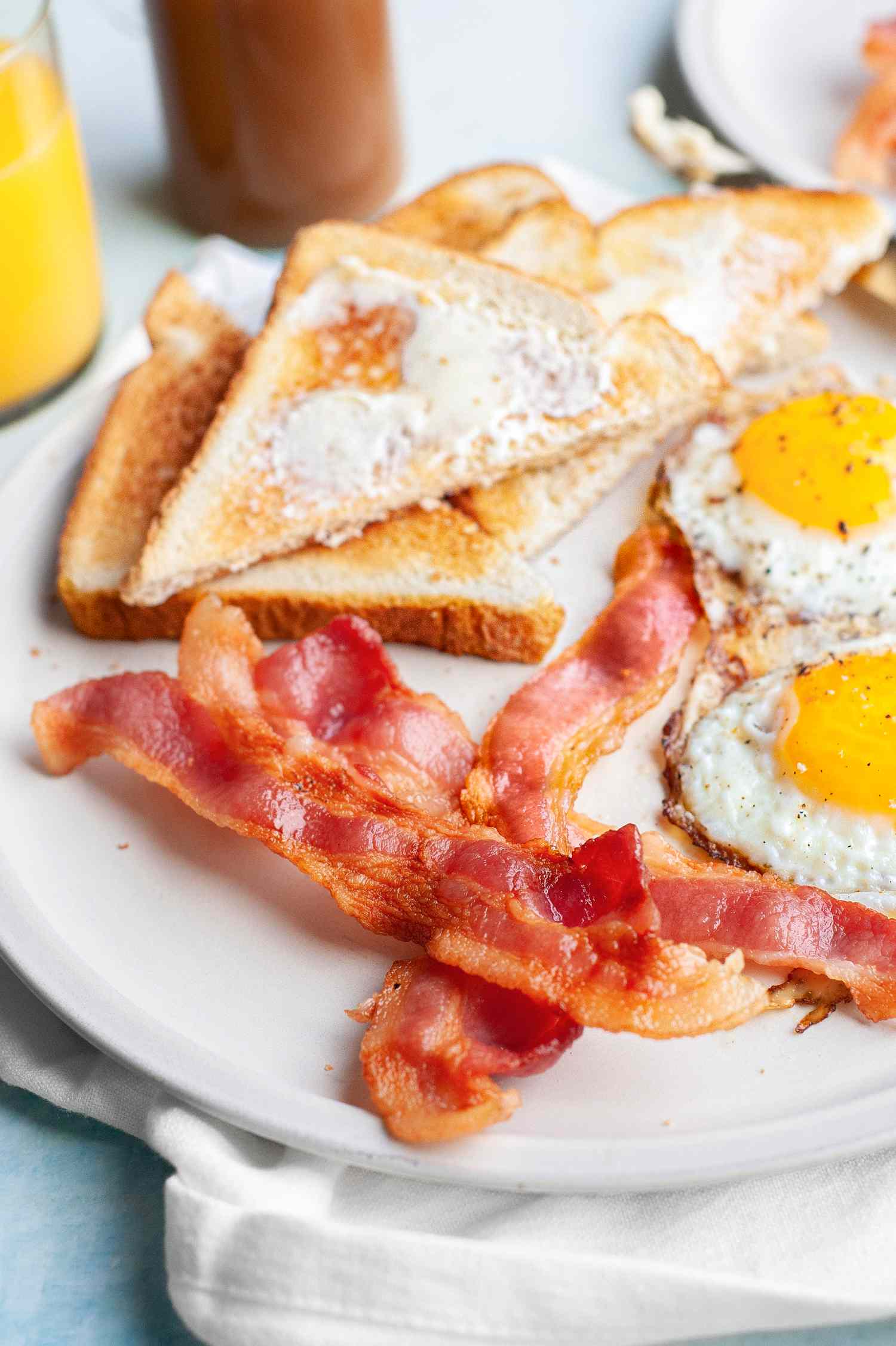 Plate of Microwave Bacon with Sunny Side Eggs and Slices of Buttered Toast, Surrounded by a Glass of Orange Juice, Mug of Coffee, and a Plate of Bacon