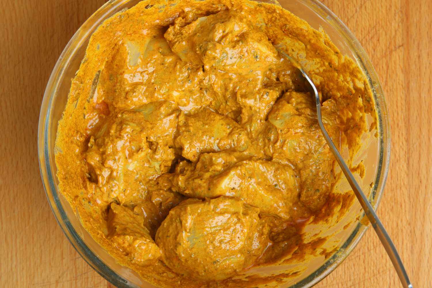 Overhead shot of a clear mixing bowl with chicken pieces getting mixed with a tandoori yogurt marinade