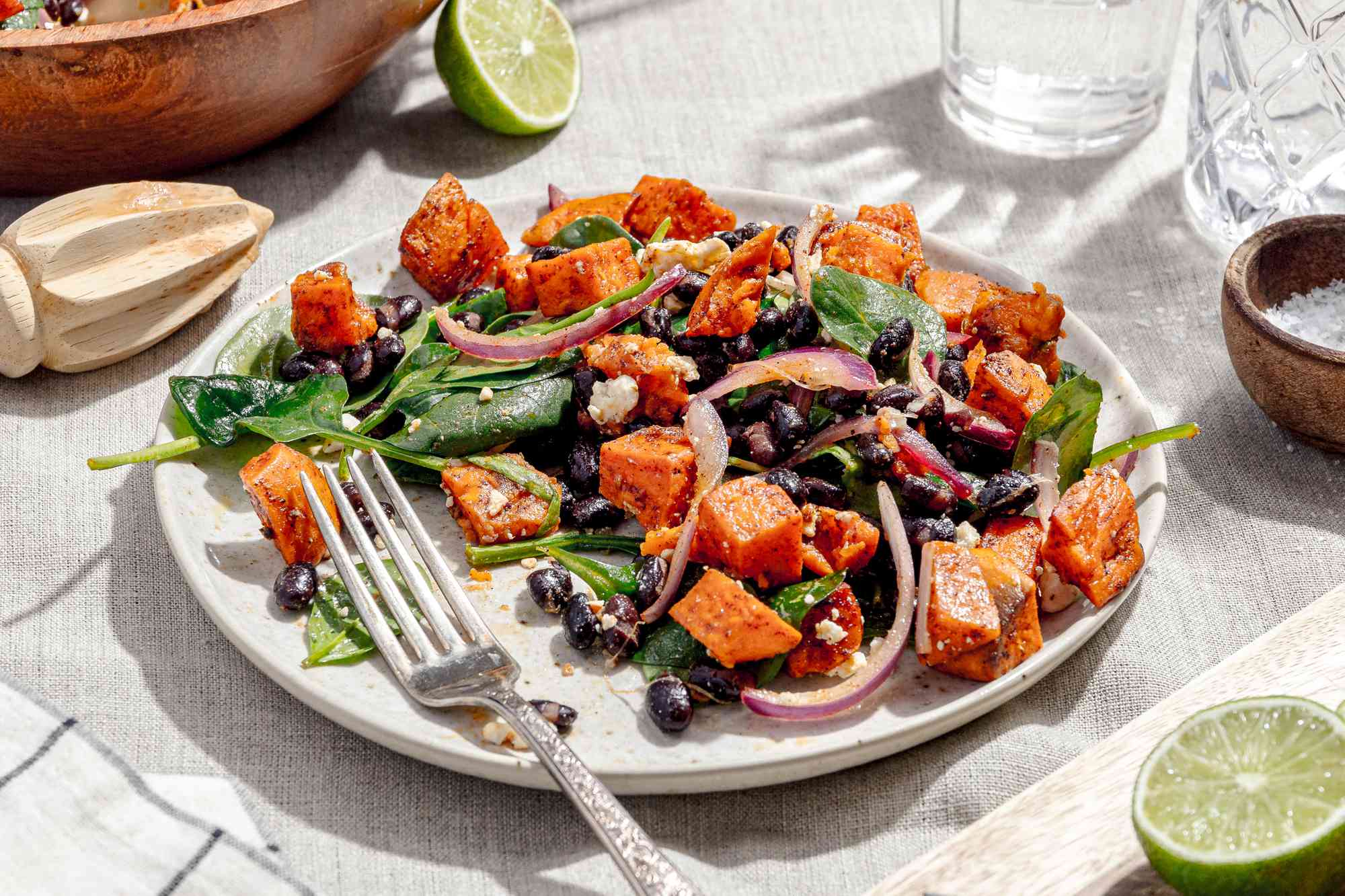 Plate of Roasted Sweet Potato Salad with a Fork, Surrounded by a Bowl with More Salad, Glasses, a Small Bowl with Salt, a Cutting Board with a Lime Half, and a Kitchen Towel