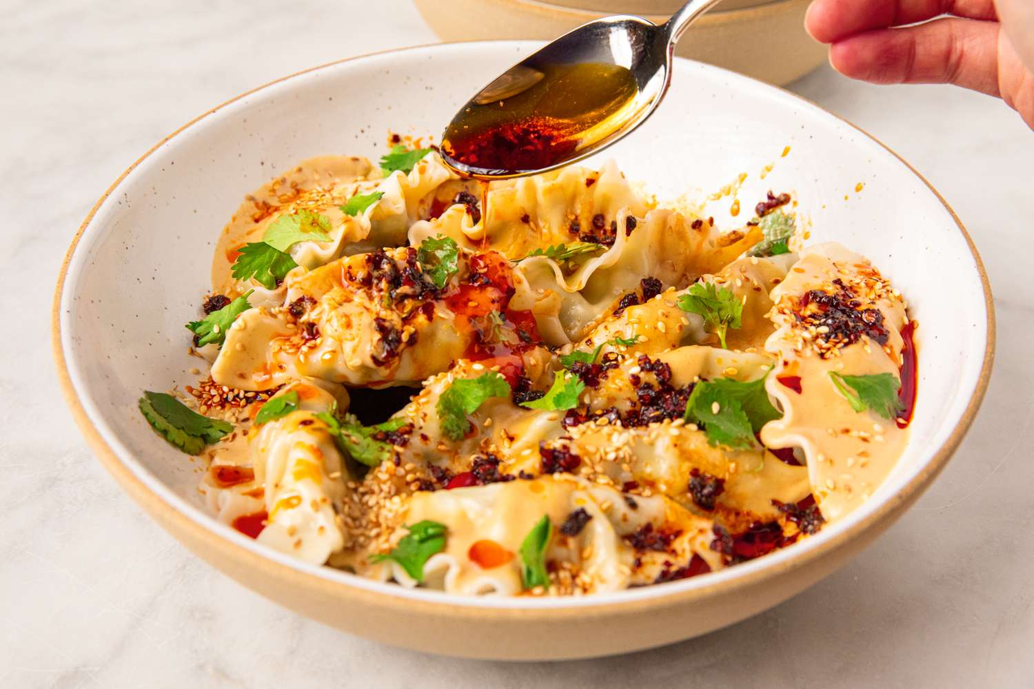 Dumplings on a plate topped with peanutchili sauce and garnished with sesame seeds and herbs with a hand pouring chili oil from a spoon