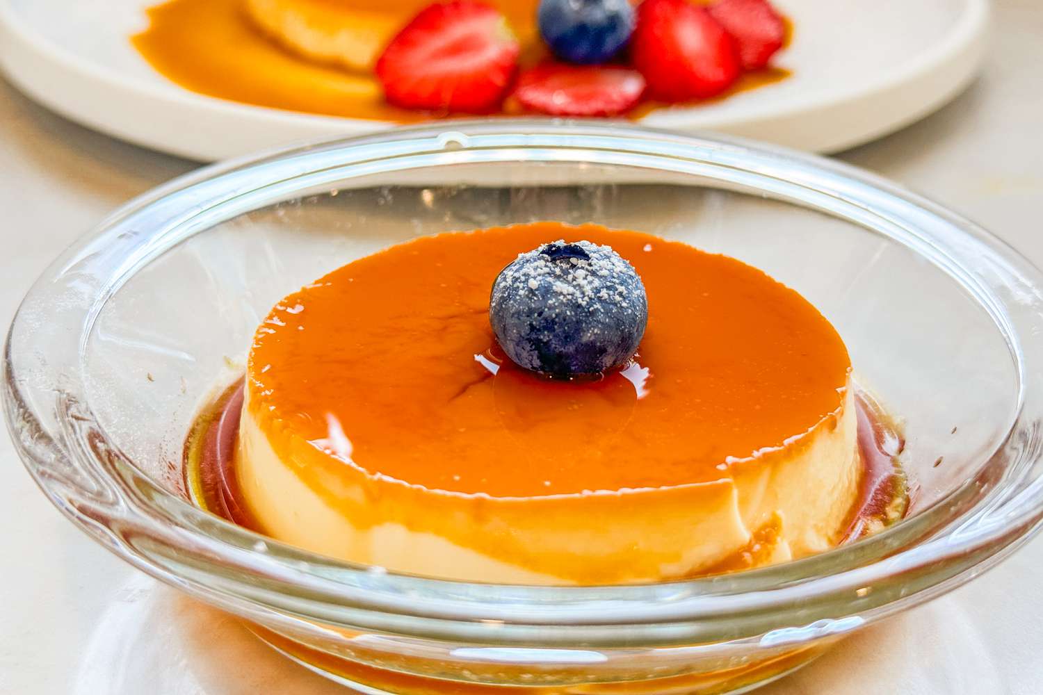 A flan in a clear bowl topped with a fresh blueberry and powdered sugar