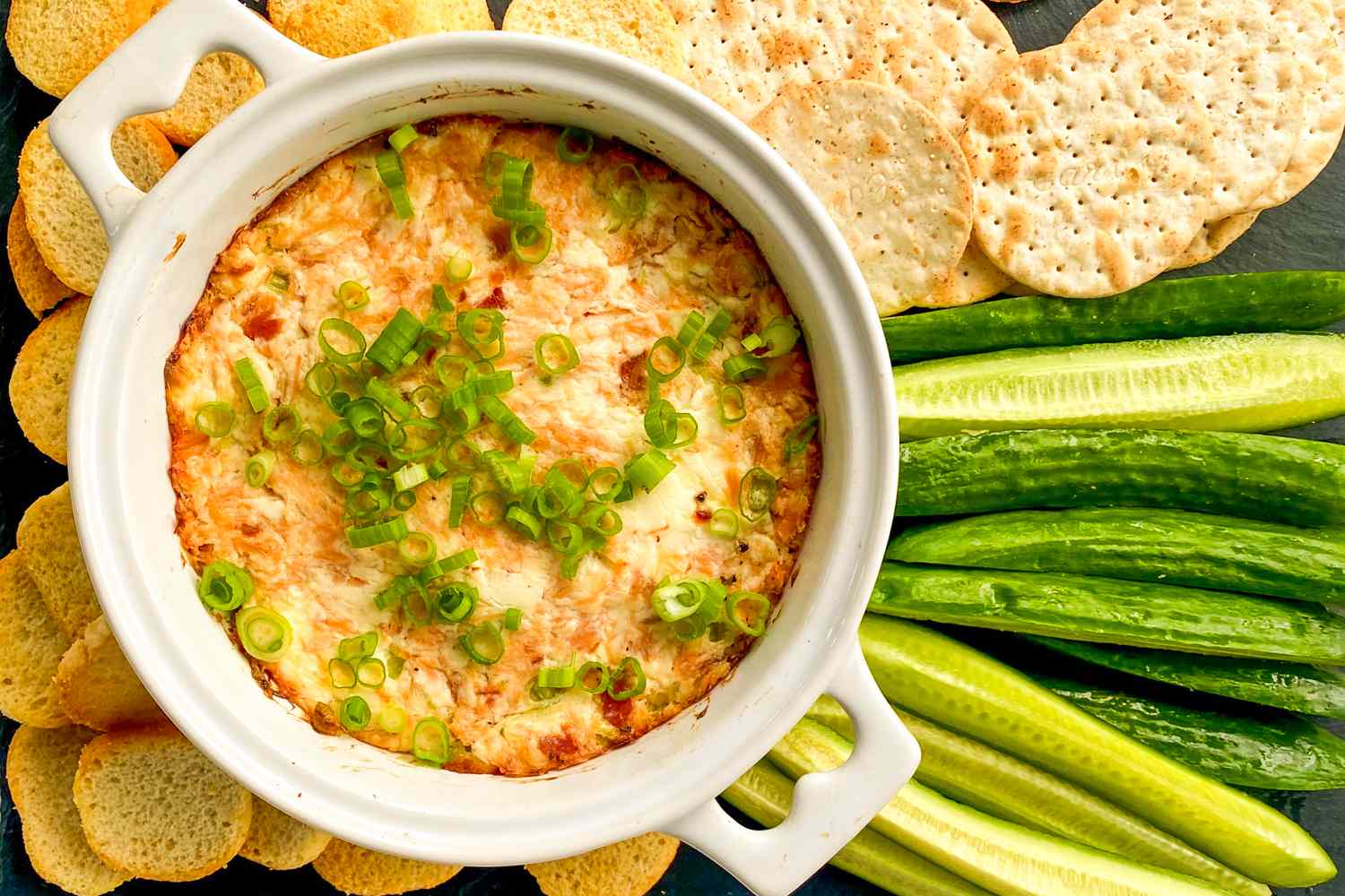 Overhead view of a white bowl of smoked salmon dip topped with green onions and surrounded by crackers, baguette slices and sliced cucumbers