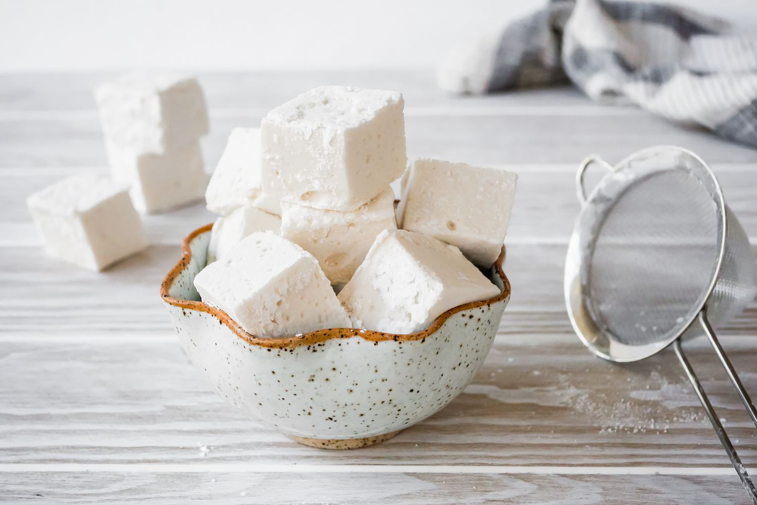 Bowl of Vegan Marshmallows with More Marshmallows in the Background. Next to the Bowl, a Small Sieve. 