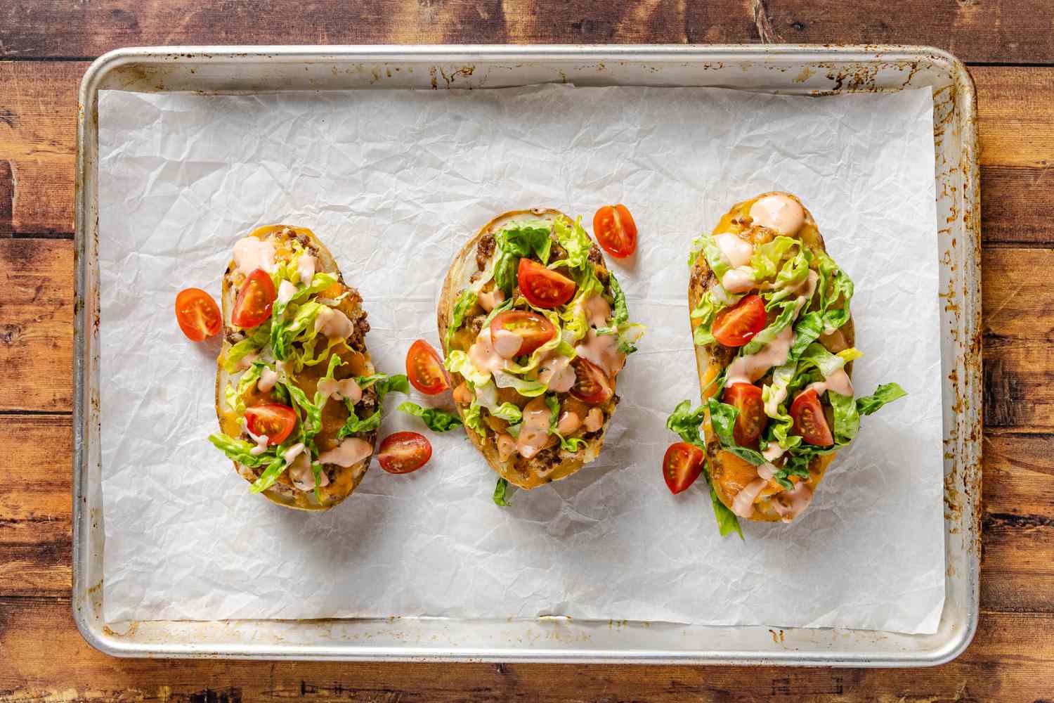cheeseburger stuffed baked potatoes topped with shredded lettuce and sliced tomatoes on a parchment paper-lined baking sheet