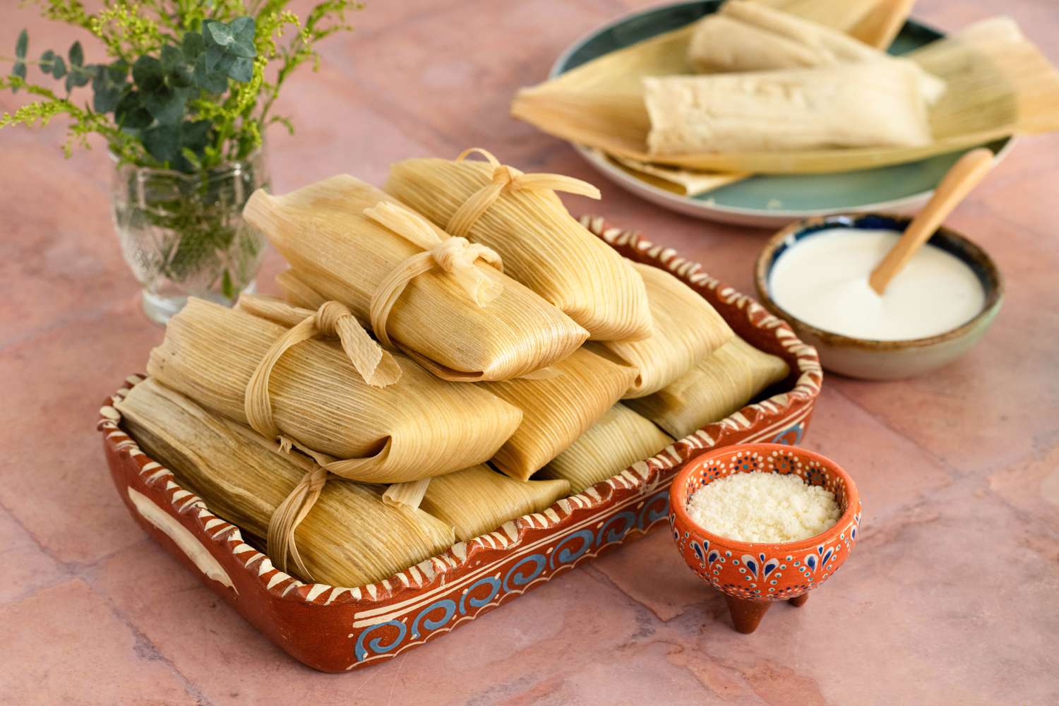 Platter of Chicken Tamales with Chile Verde Next to a Small Bowl of Crema, a Small Bowl of Cotija Cheese, a Glass of Water, Small Vase, and a Plate with an Opened Tamale