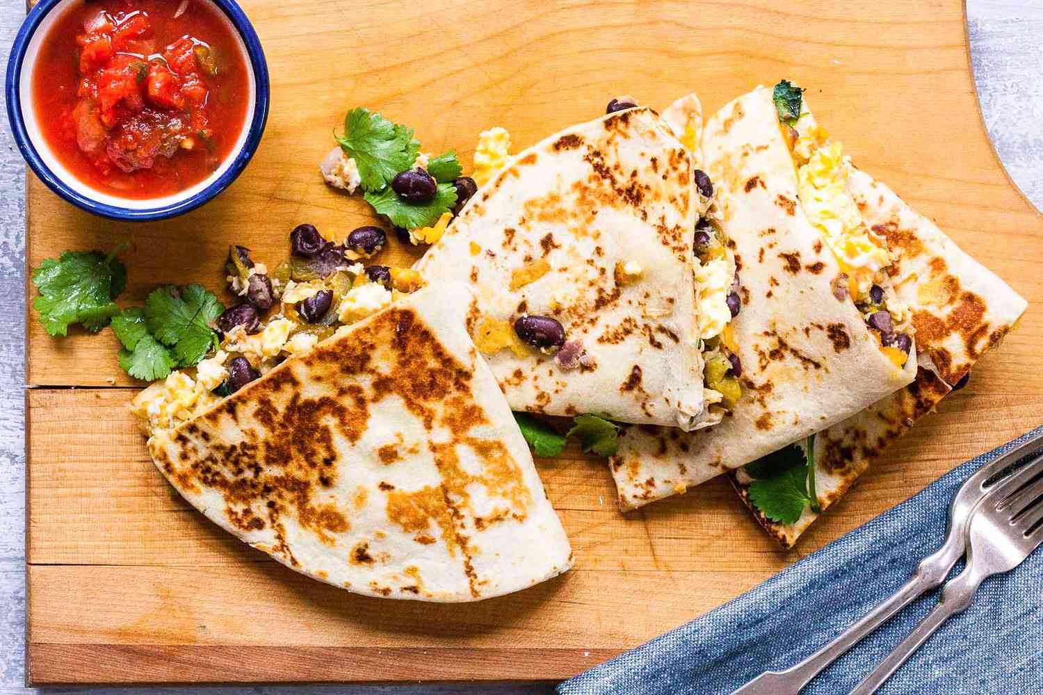 Baked simple breakfast quesadillas on a wood cutting board with cilantro, black beans and scrambled eggs visible inside. The quesadillas are partially stacked and two forks and a blue linen are below and to the right. A small bowl of salsa is in the uper left corner.