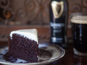 A Slice of Chocolate Guinness Cake on a Plate and in the Background, a Bottle of Guinness and More in a Glass 