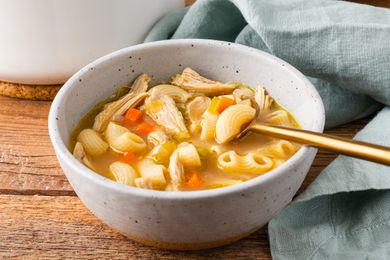 Bowl of soup with pasta shredded chicken and vegetables with a spoon and cloth napkin nearby