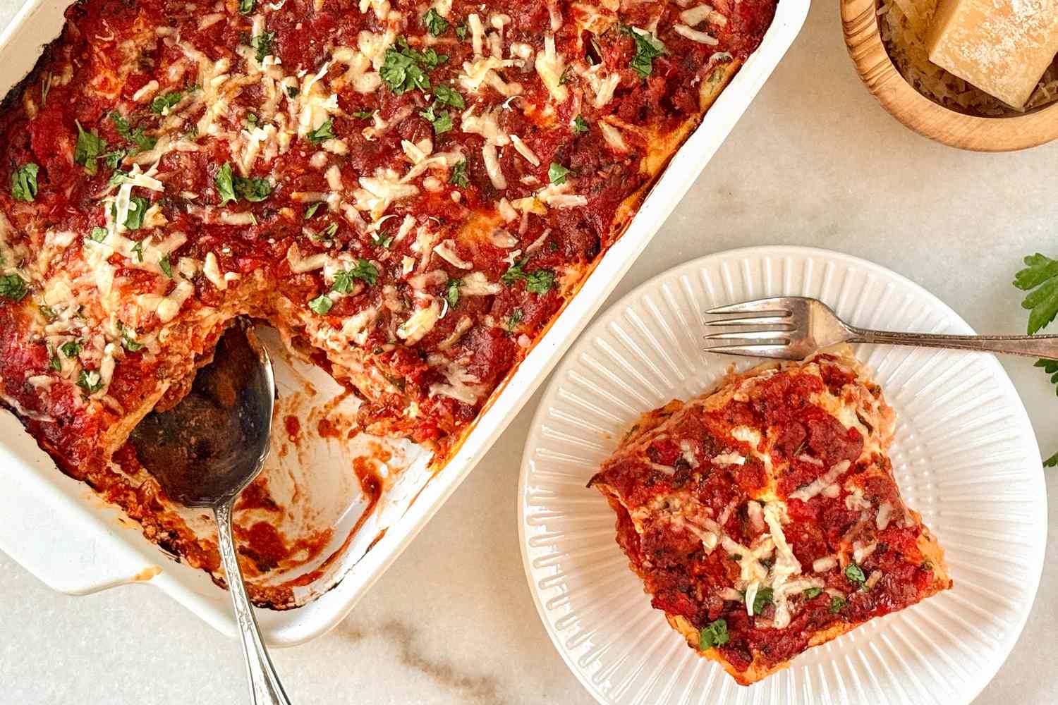 A casserole dish with lasagna and a serving on a plate with a fork accompanied by a wedge of Parmesan cheese in a bowl