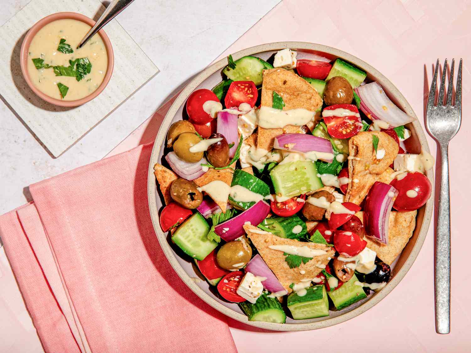 Bowl of salad topped with hummus salad dressing, and in the surroundings, a bowl of hummus salad dressing on a white coaster and a pink table napkin