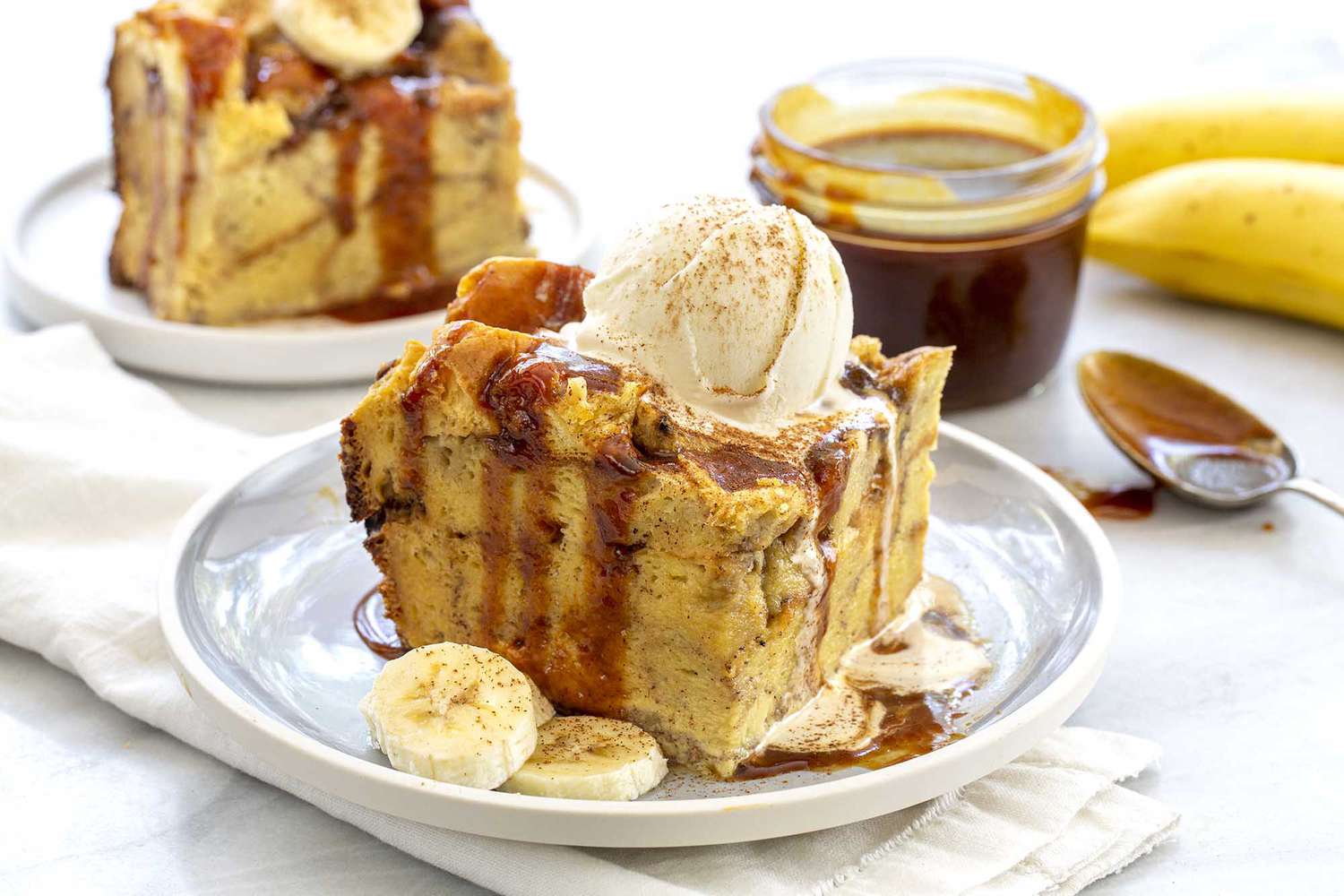 Slice of the best slow cooker banana bread pudding topped with a scoop of ice cream and drizzeld with caramel sauce on a gray plate. A jar of caramel sauce is off to the side.