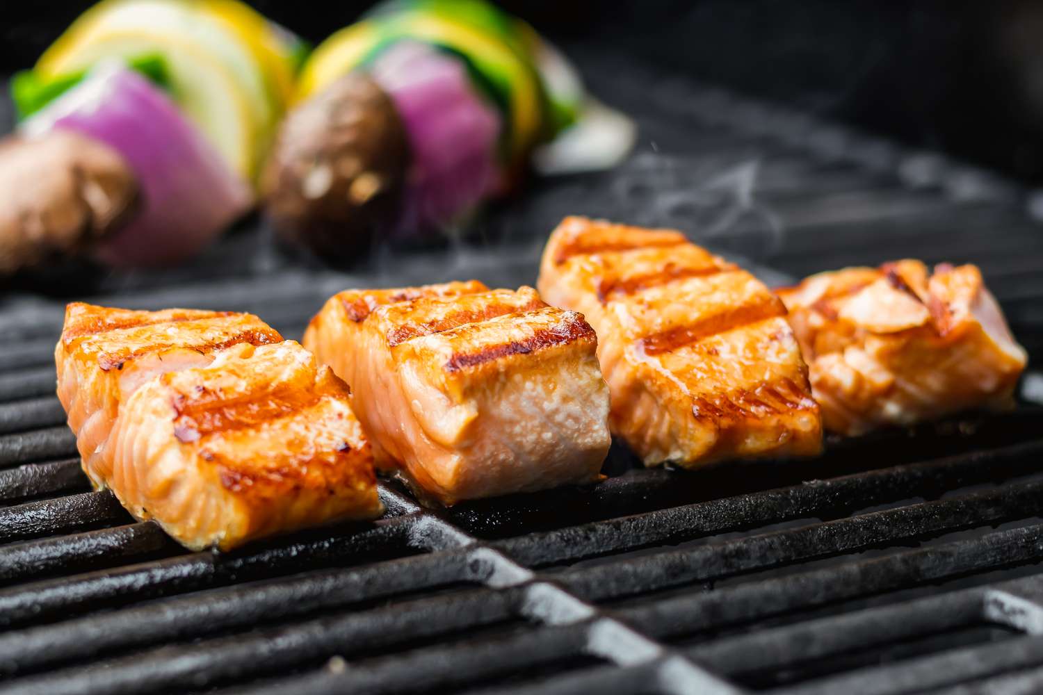 Four salmon fillets on a grill with veggie skewers cooking in the background