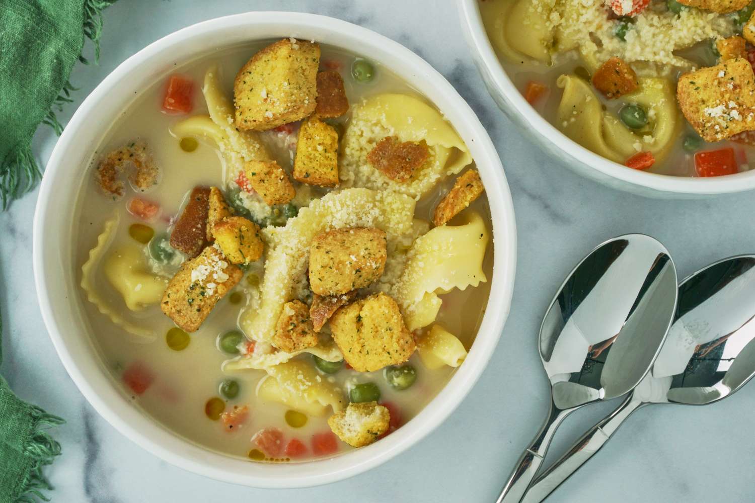 Bowls of tortellini soup topped with croutons on a table setting with spoons and a napkin