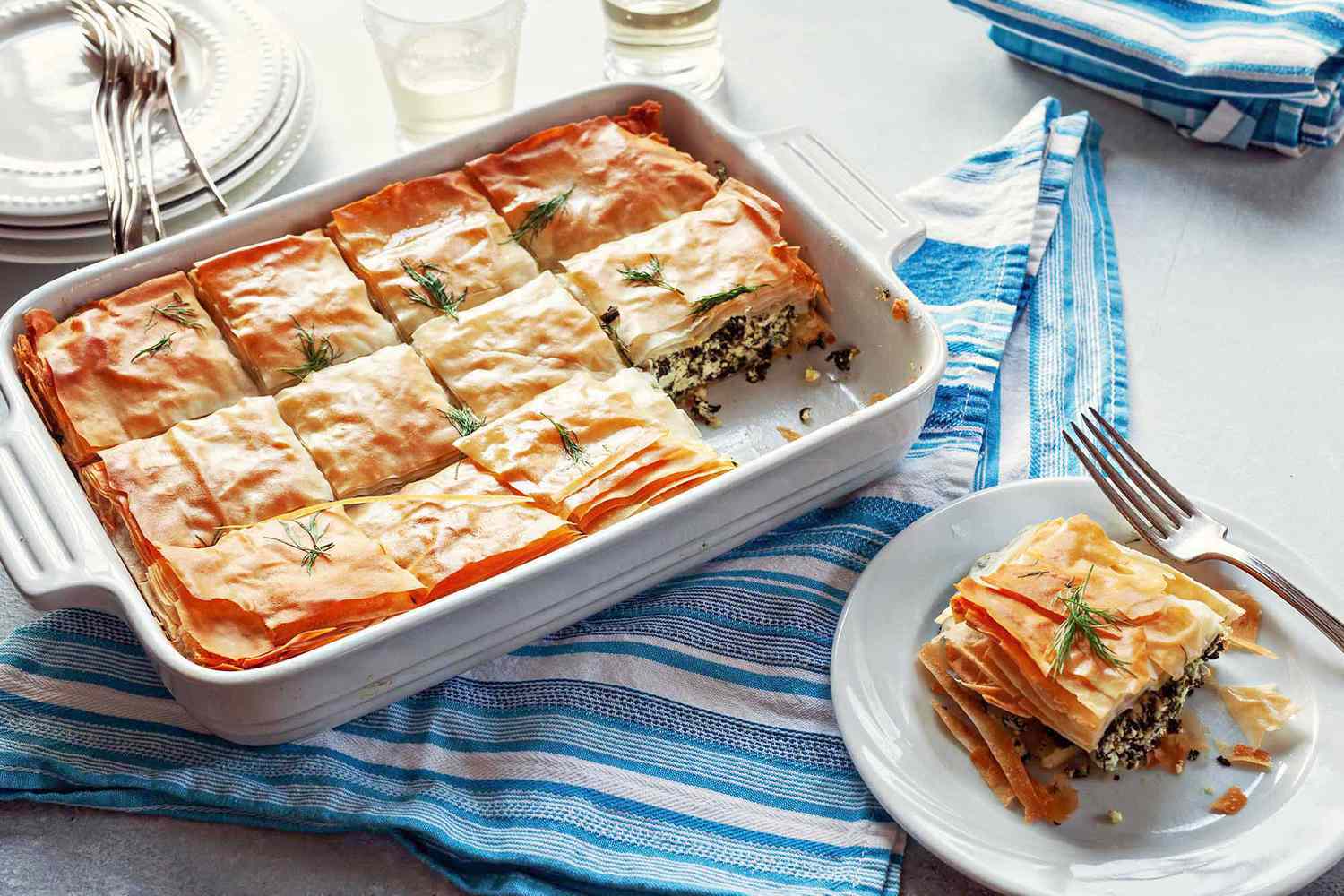 Slice of Greek Spinach Pie on a white plate, with a fork, a glass of white wine, and the casserole dish in the background,