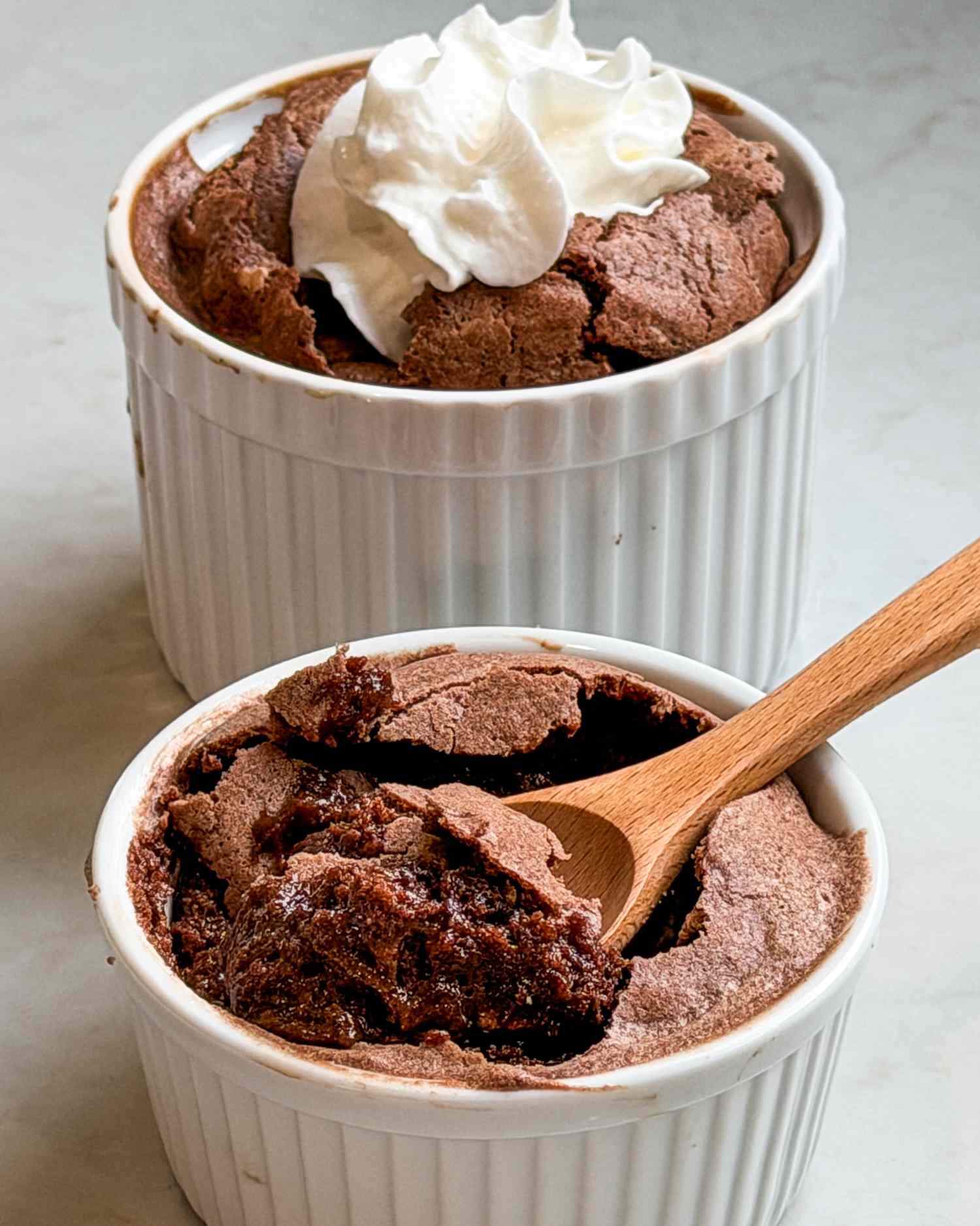 Two ramekins of brownie pudding one topped with whipped cream and the other with a wooden spoon digging in