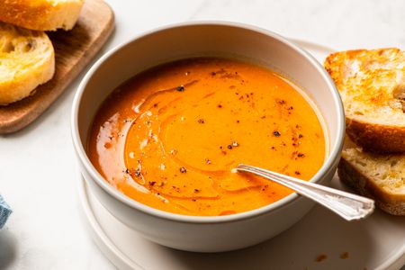 A bowl of tomato and red lentil soup with a spoon served with bread slices on the side