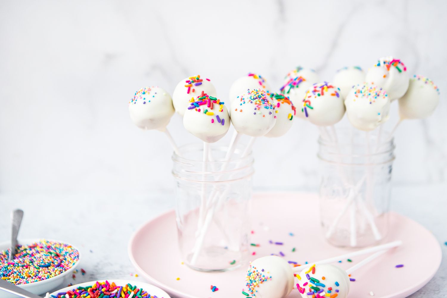 Sprinkled Cake Pops in Jars on a Plate Next to Two Small Plates of Sprinkles