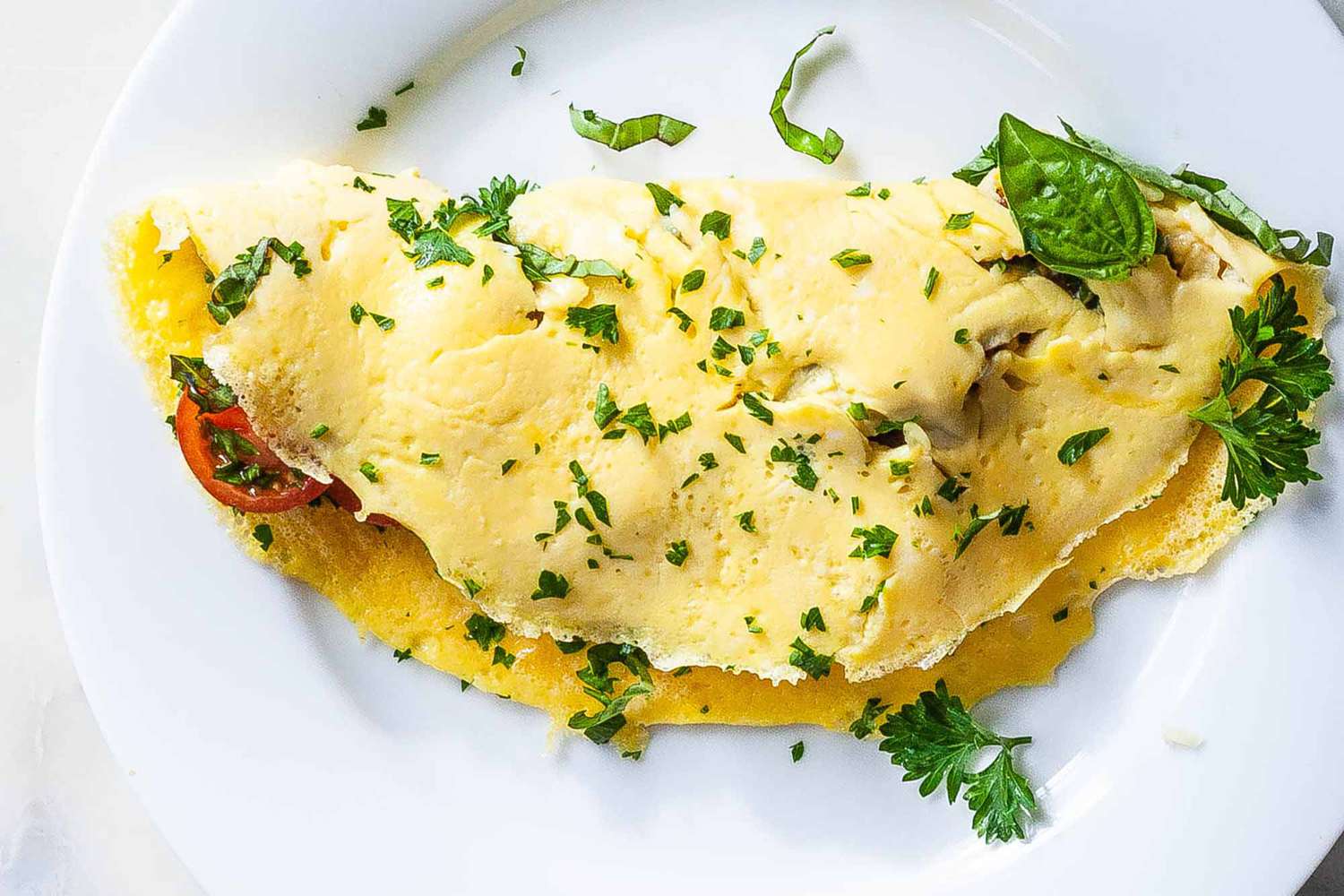 An omelette with cut cherry tomatoes inside and parsley sprinkled on top resting on a white plate