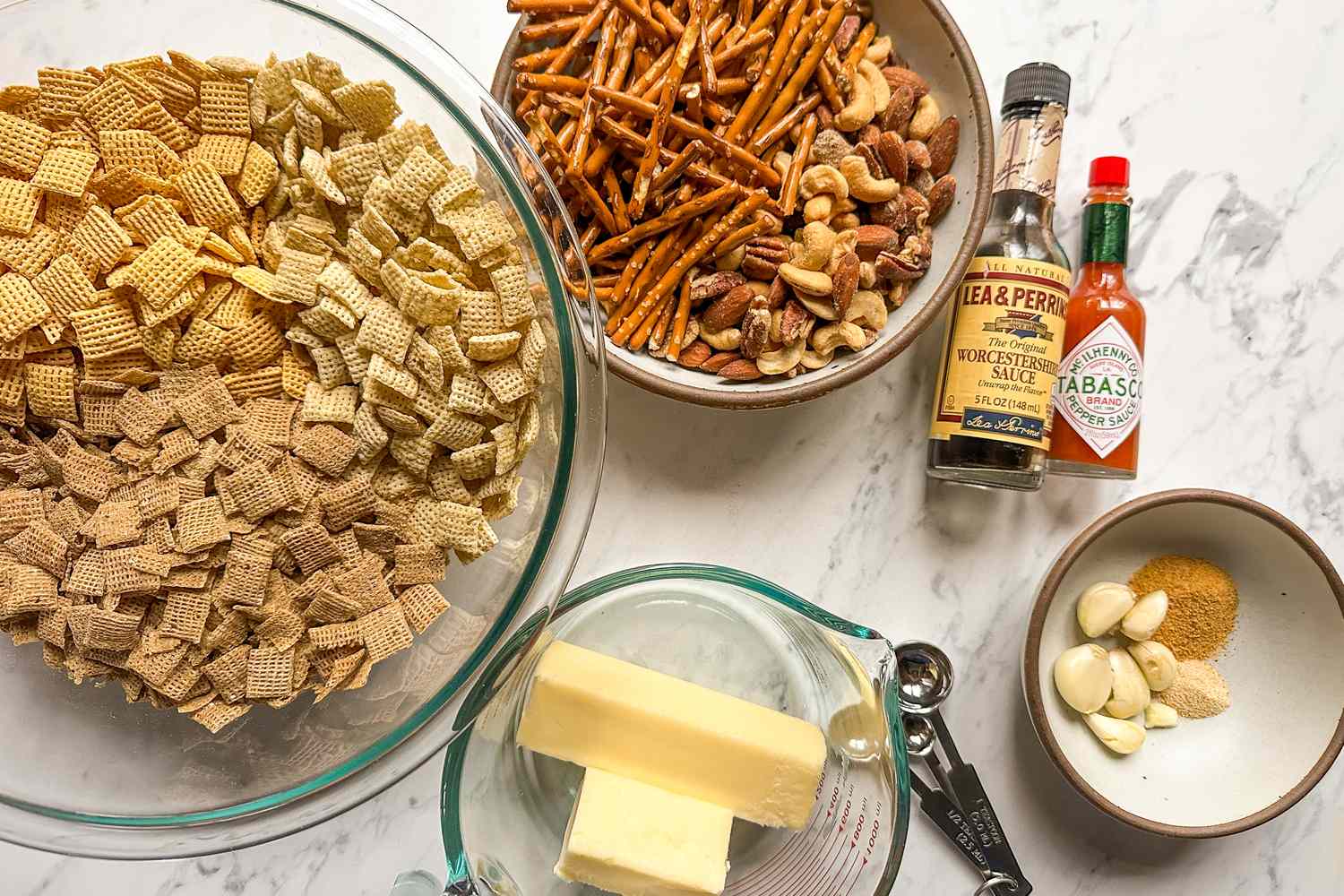 Ingredients for a snack mix including cereal pretzels nuts butter and condiments displayed on a counter