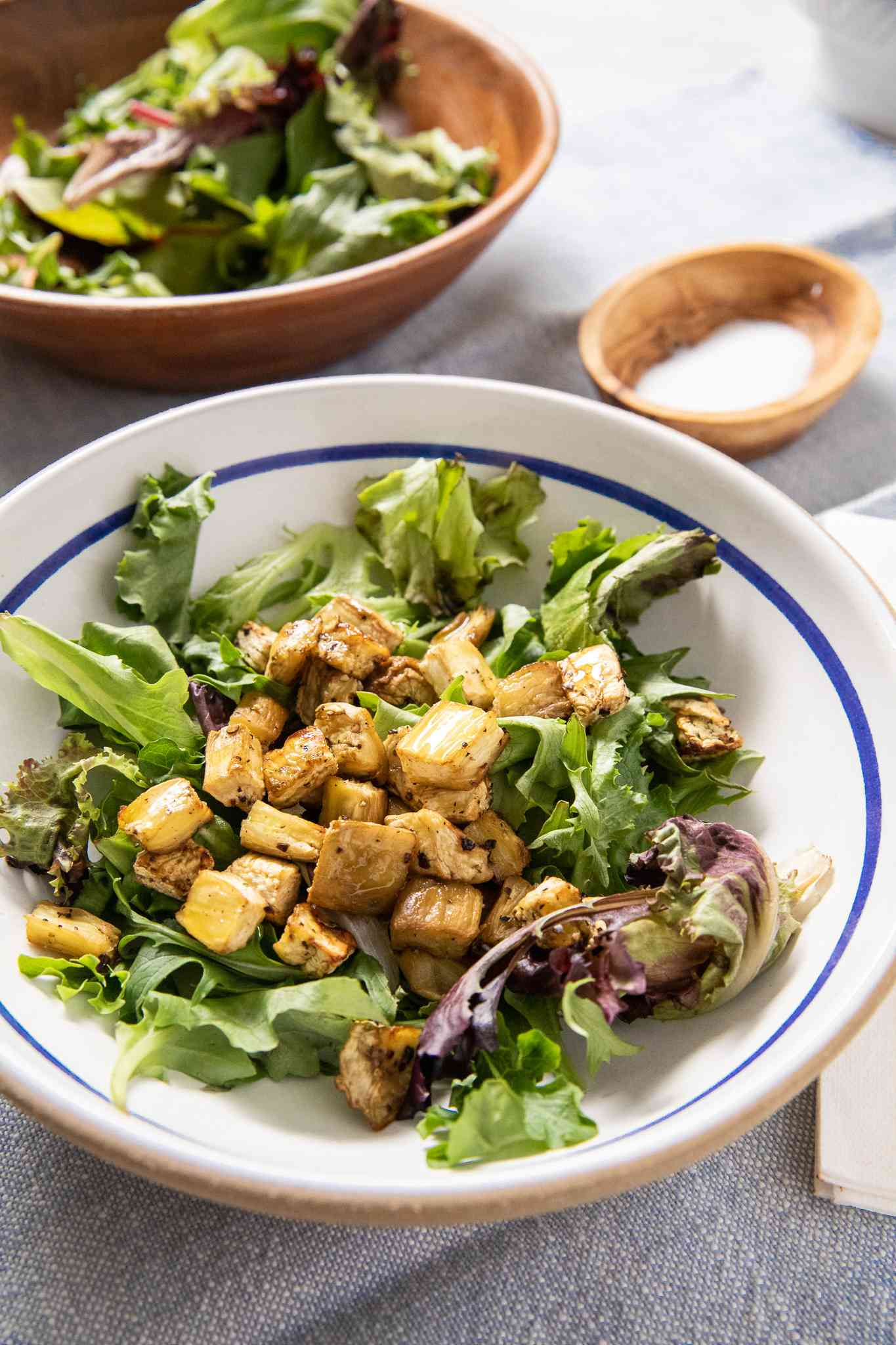 Bowl of Salad Greens Topped With Cubed Roasted Eggplant, and in the Background, Another Bowl With More Salad Greens and a Small Bowl of Salt
