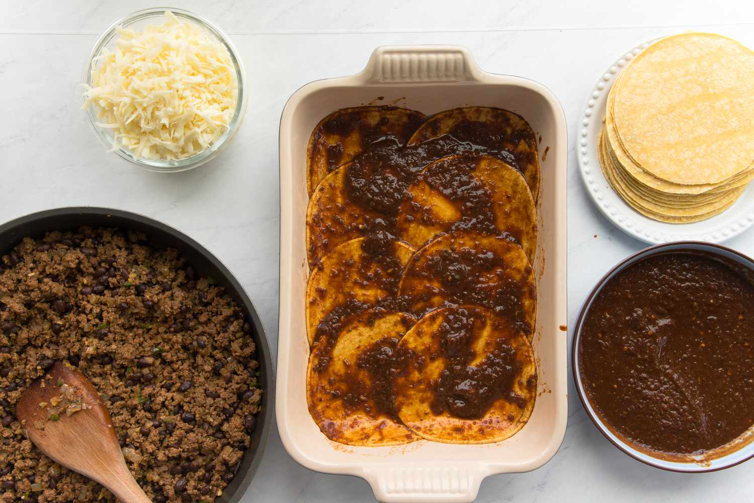 Casserole Dish with Enchilada Sauce Soaked Tortillas Surrounded Enchilada Ingredients (a Plate of Tortillas, Bowl of Sauce, Bowl of Shredded Chicken, and Bowl of Shredded Cheese)